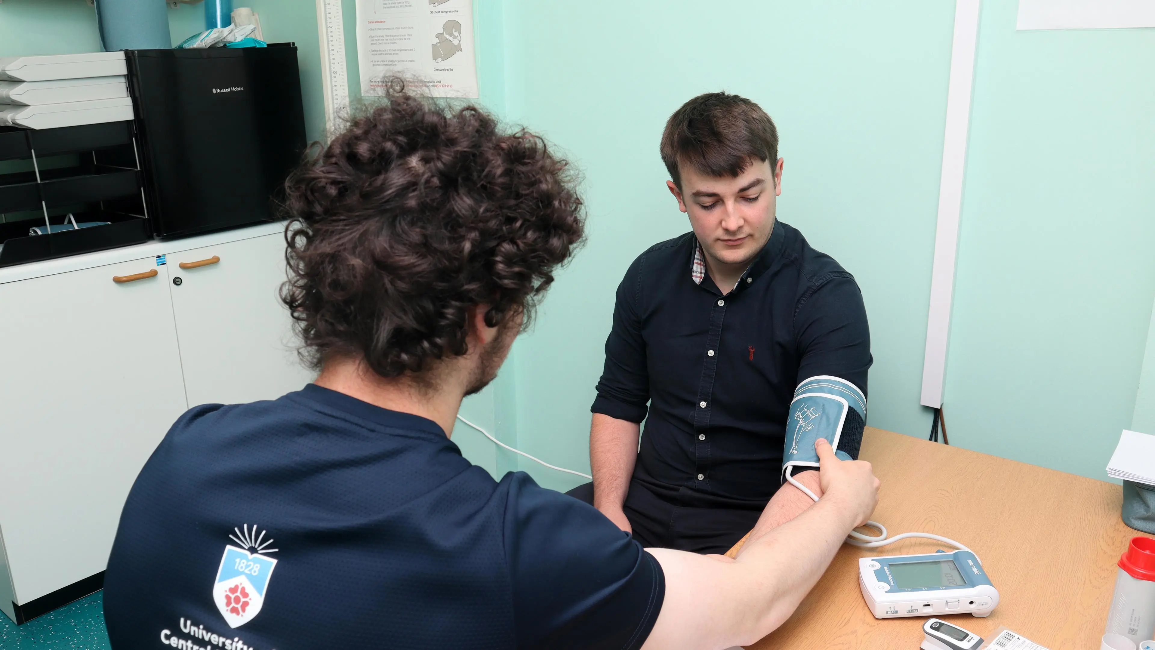 Two male students are sat down demonstrating how to use a blood pressure monitor.