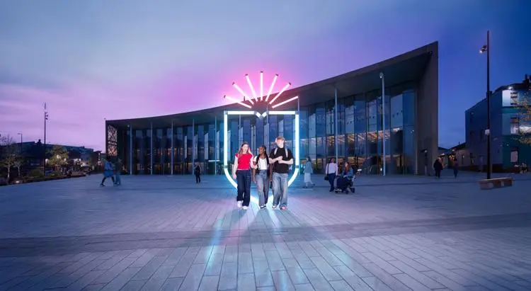 Students outside the student centre at sunset with a neon shield behind them