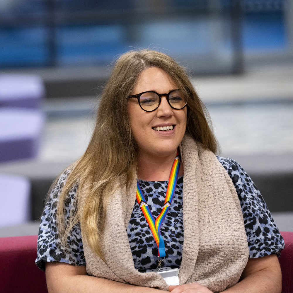 harriet tyler from the people team sat on a burgundy sofa with a blue leopard print top on, glasses and a rainbow lanyard