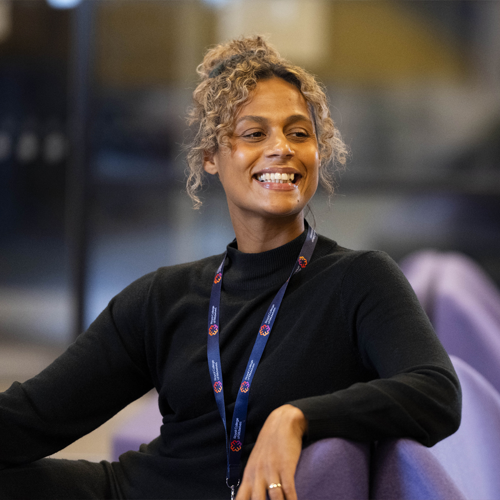 midwifery lecturer kimberley olorenshaw sat on a purple sofa smiling in a black turtle neck jumper and lanyard