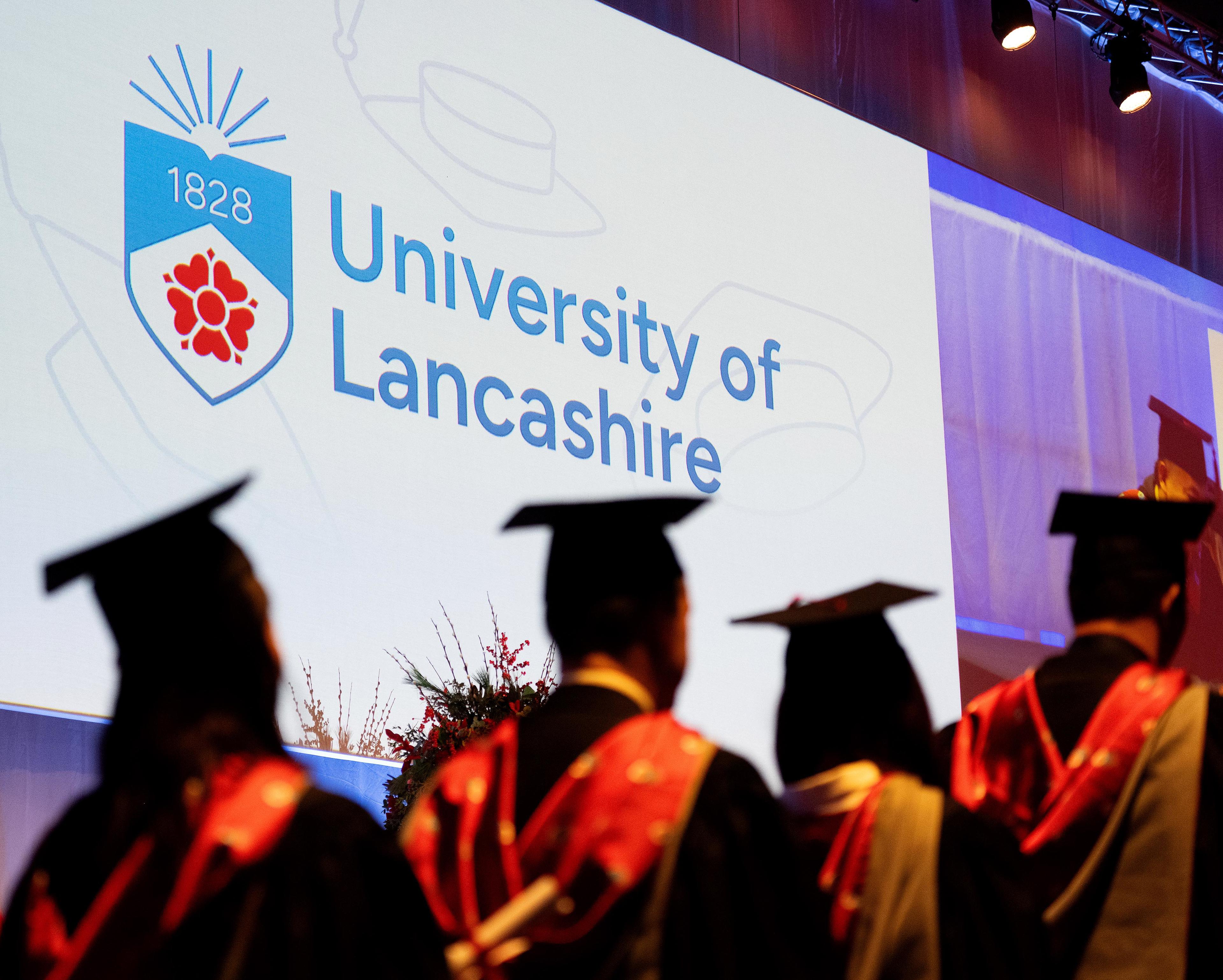 Four graduates in academic robes with a large logo on a big screen